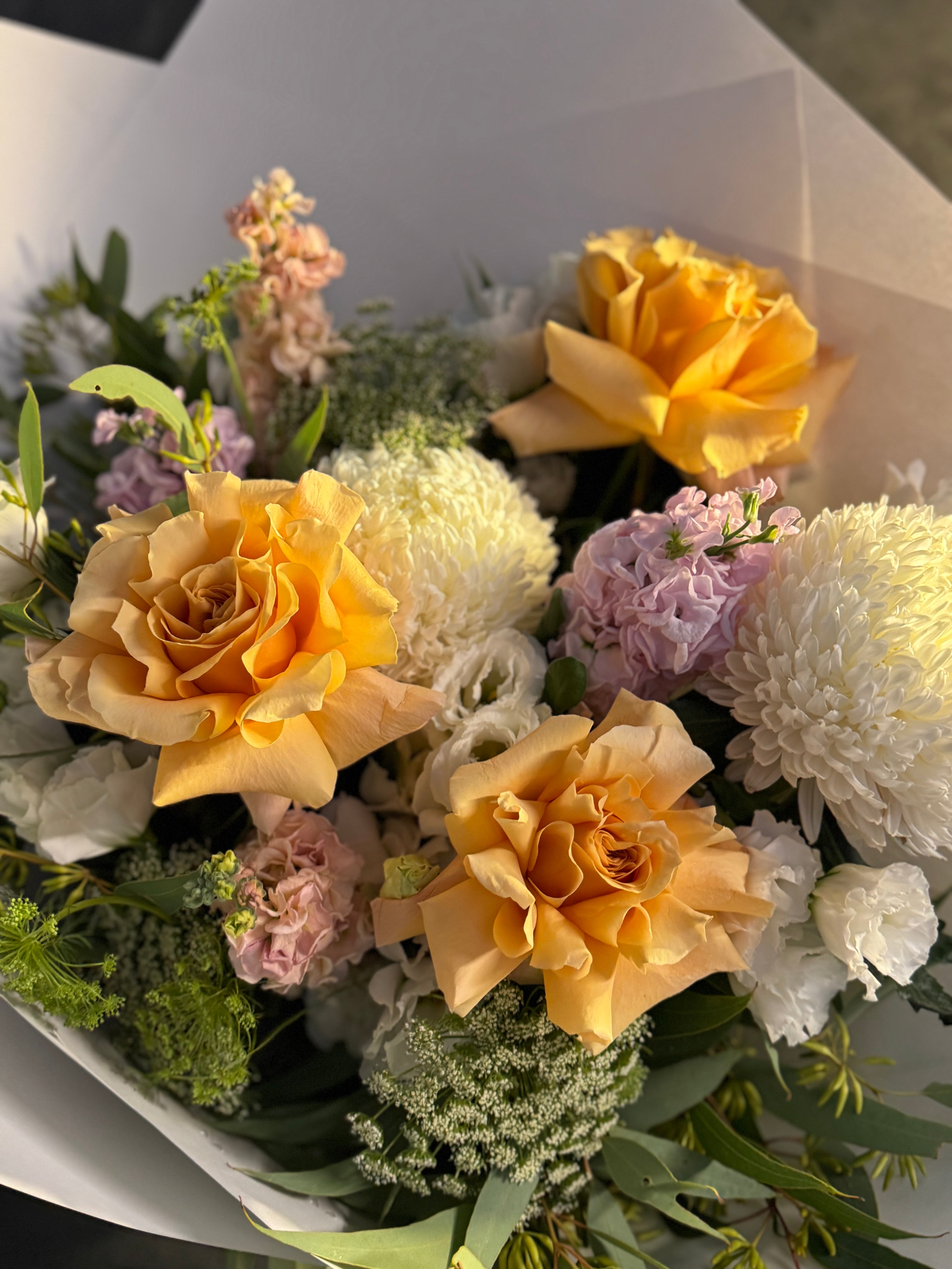 Bouquet of yellow roses, white chrysanthemums, and light pink flowers with greenery, wrapped in white paper.